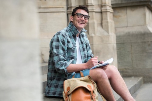 A student wearing shorts and a chequered shirt smiles at the camera with a notepad and pen in hand.